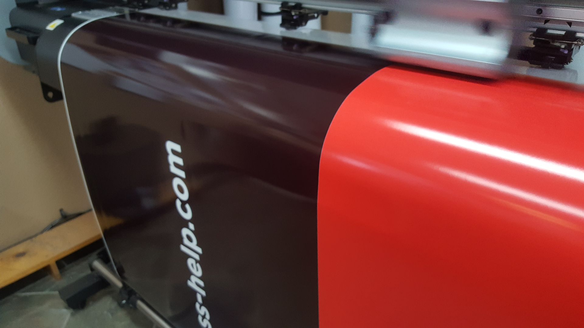 Large format printer printing a red vinyl sheet, next to a black machine with text.