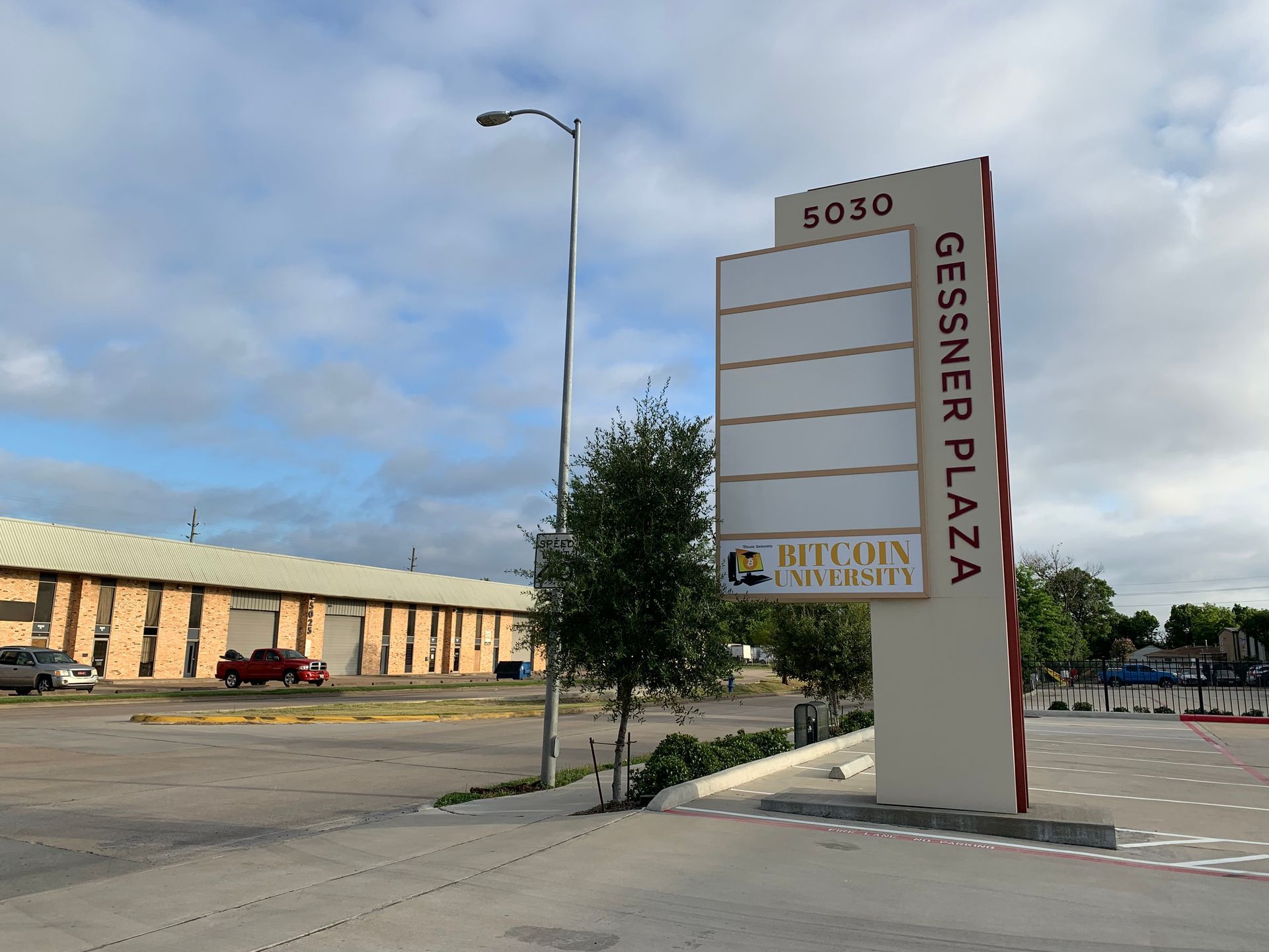 Gessner Plaza sign with blank panels, on a beige pole. A brick building and cars are in the background.