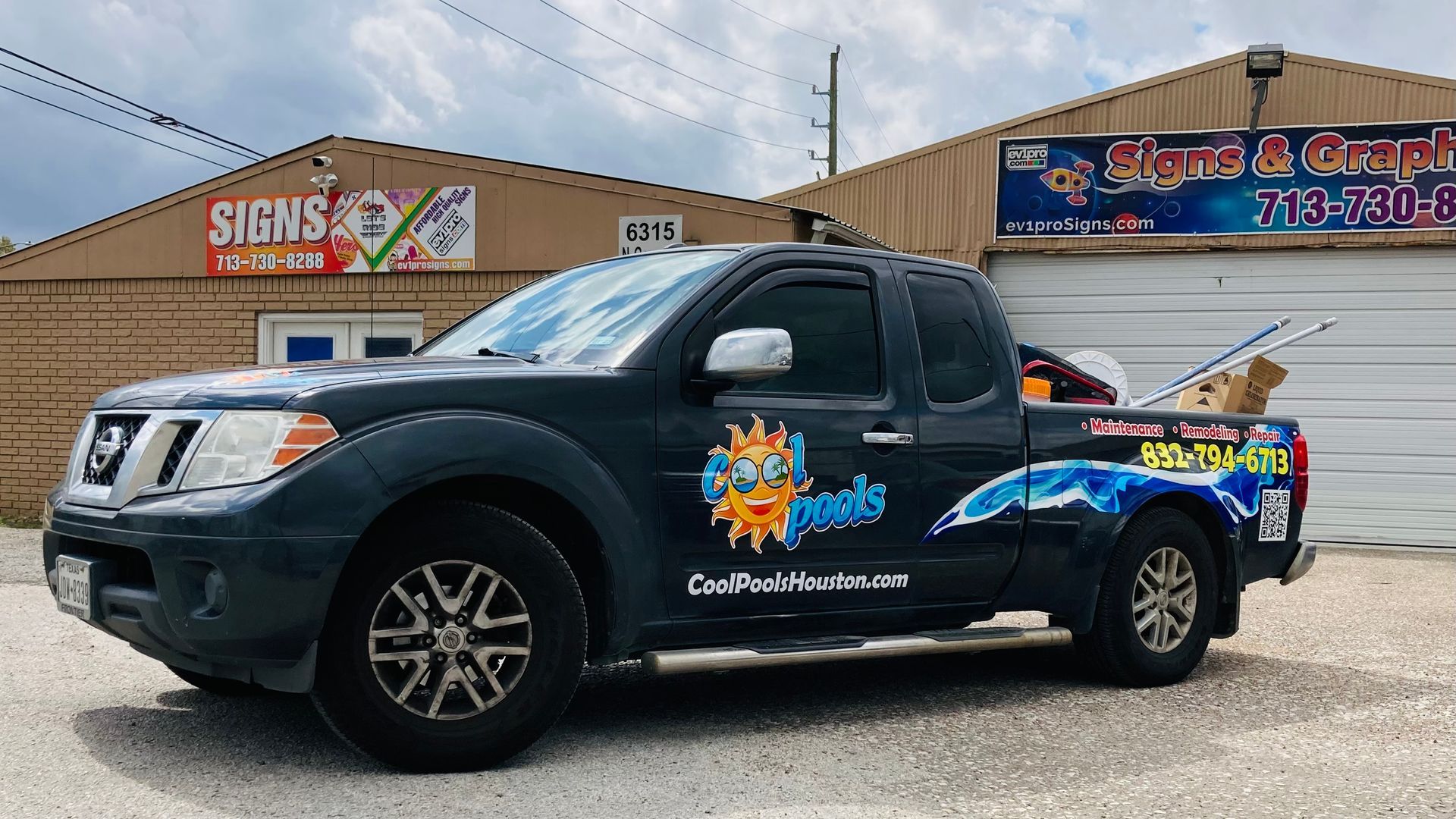 Dark pickup truck with company logo parked outside a sign shop. Building has signage and a garage door.