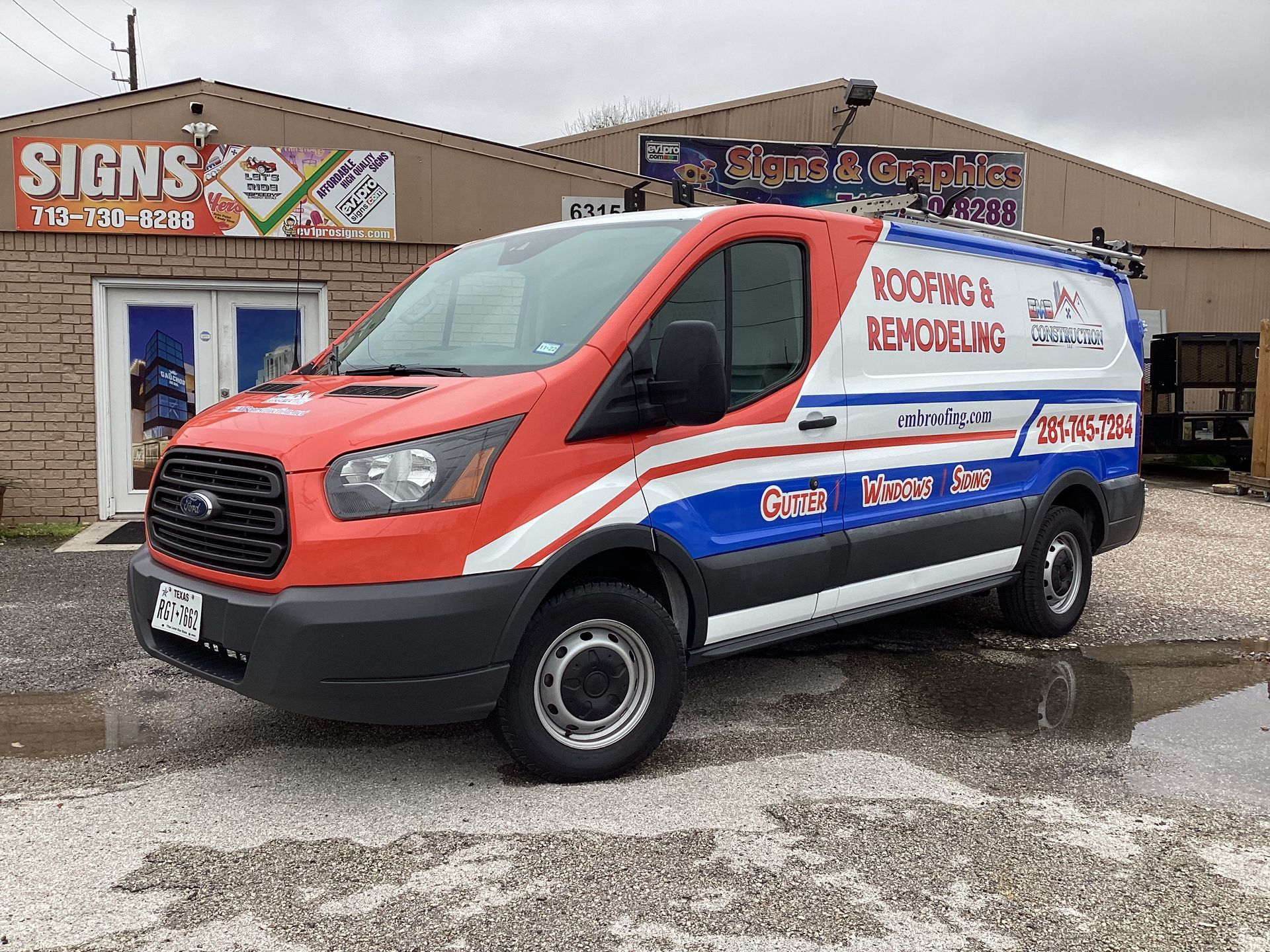 Red, white, and blue van parked outside a building; the van has roofing and remodeling company branding.