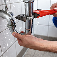 Plumber using a wrench to fix a leaky pipe under a sink. Water sprays around the area.