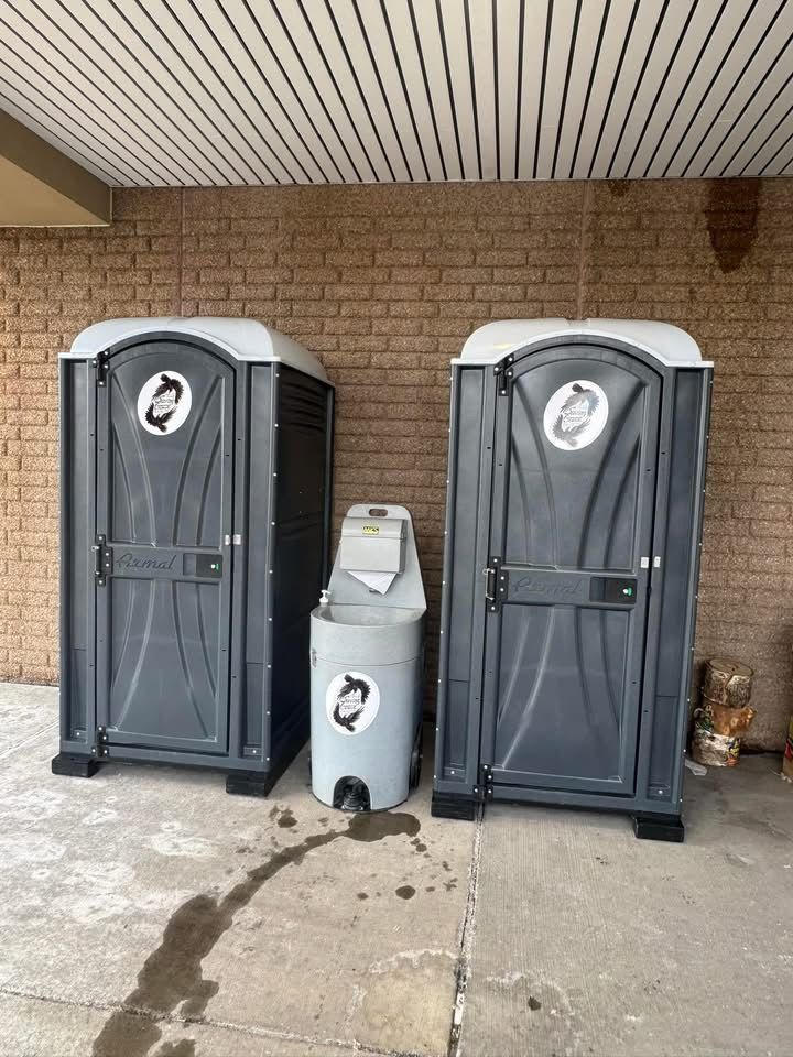 Two gray portable toilets beside a silver handwashing station under a covered brick wall.