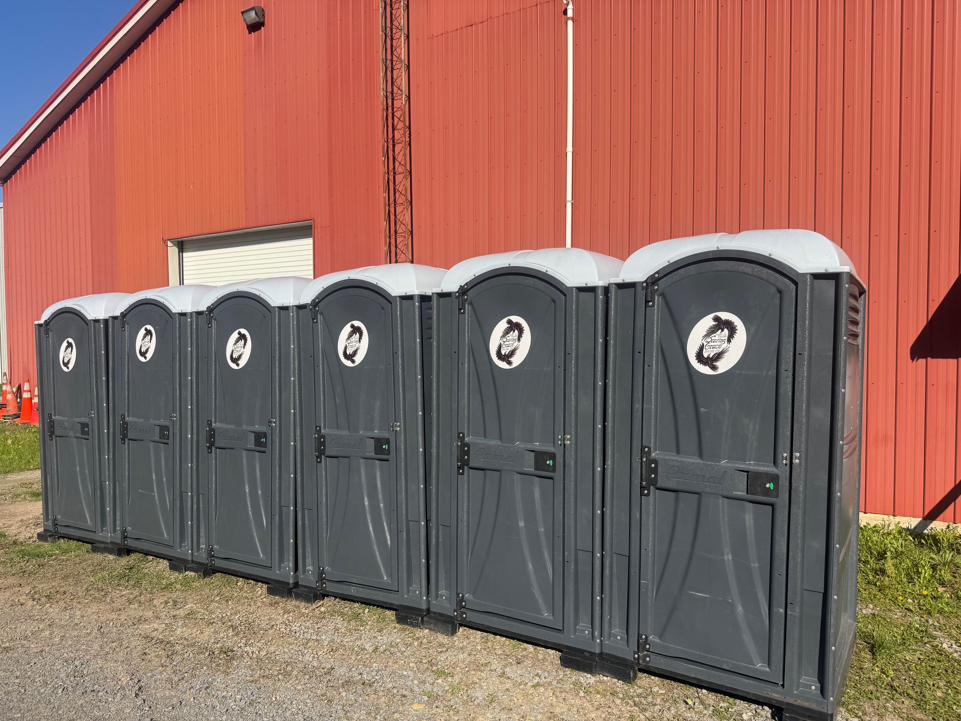 Row of dark gray portable toilets in front of a red barn on gravel