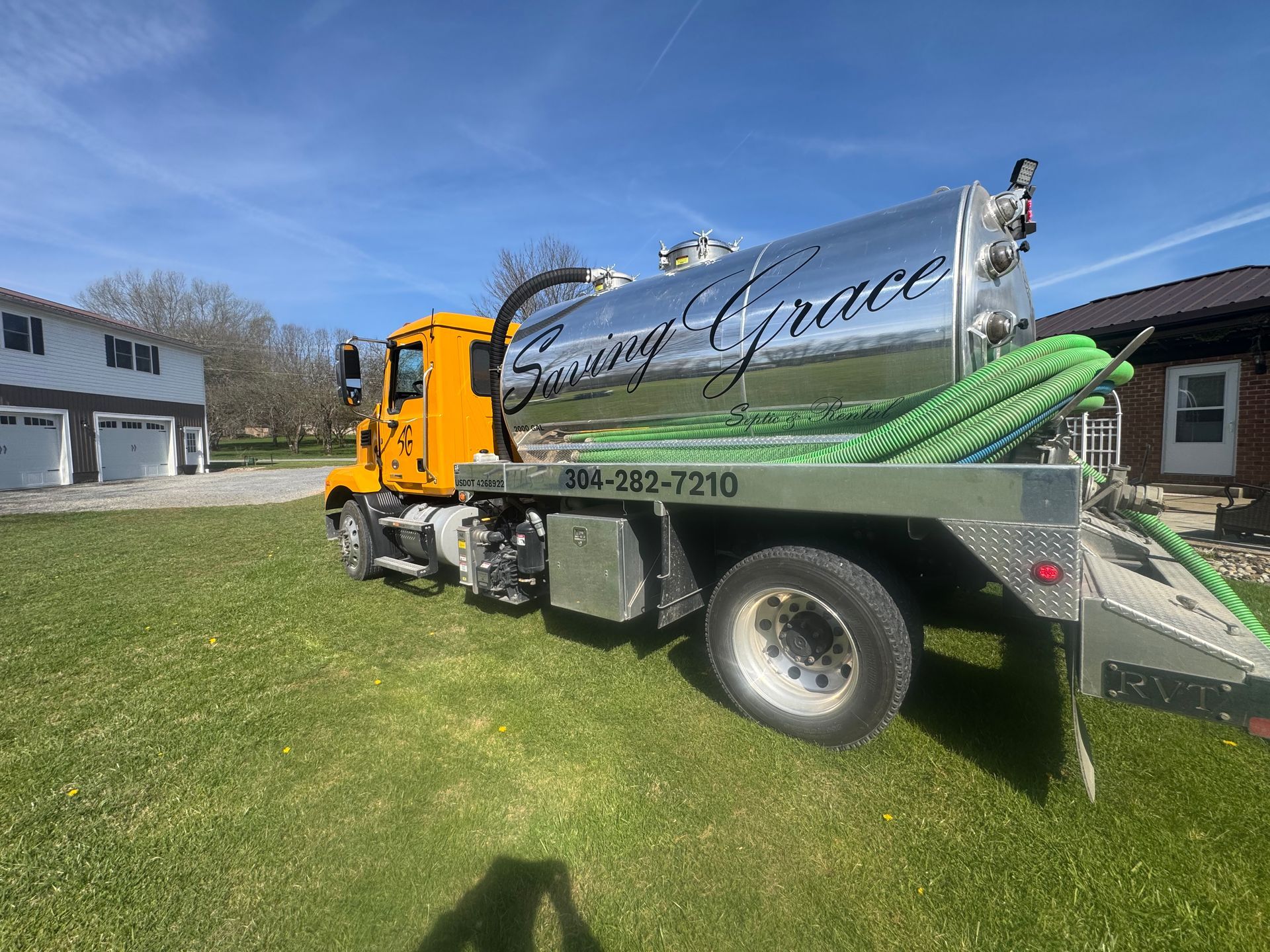 Yellow dump truck with a shiny metal tank parked on grass outside a house under a blue sky