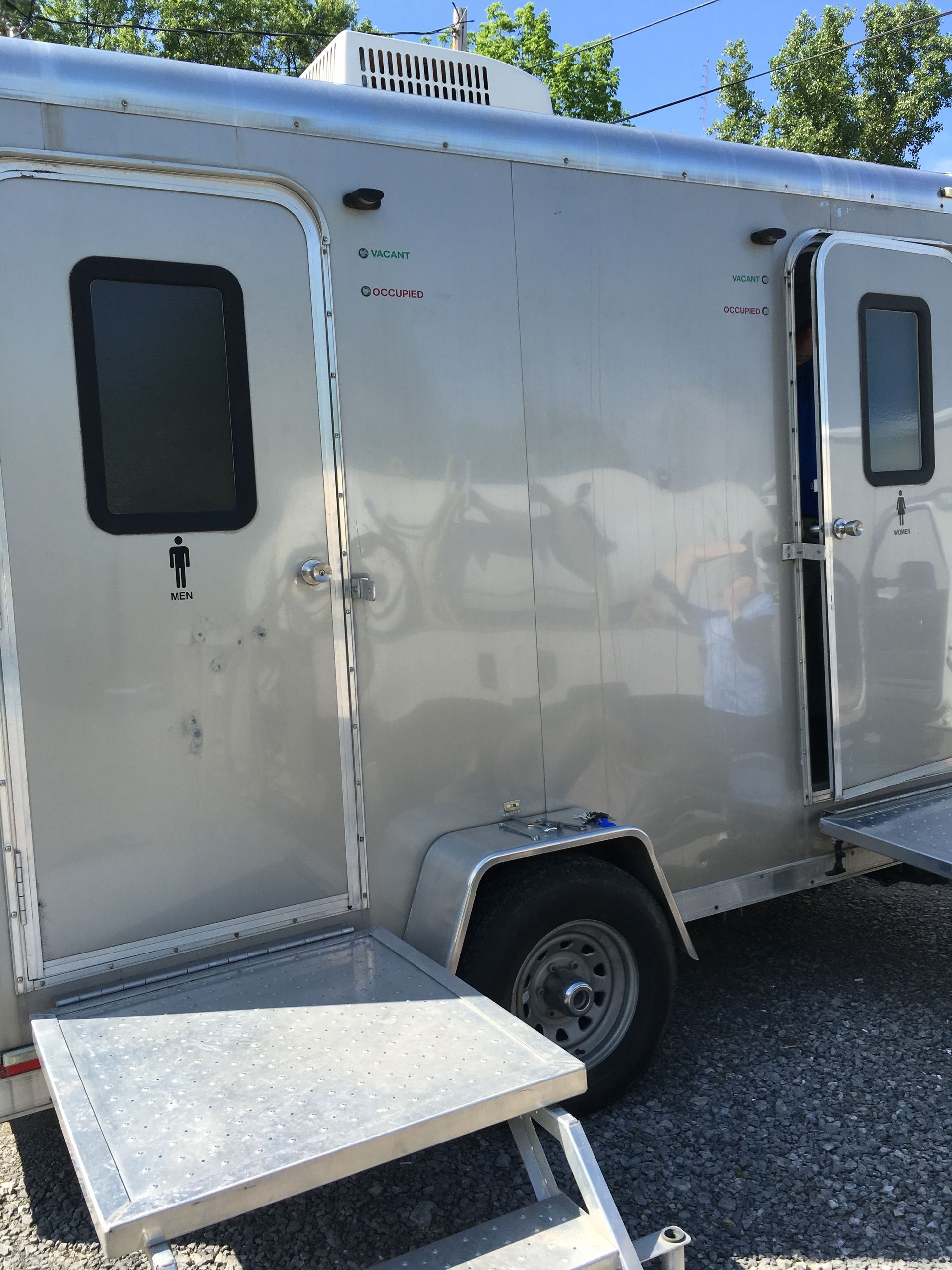 Silver utility trailer with a side door and small ramp, parked outdoors near trees and a blue sky