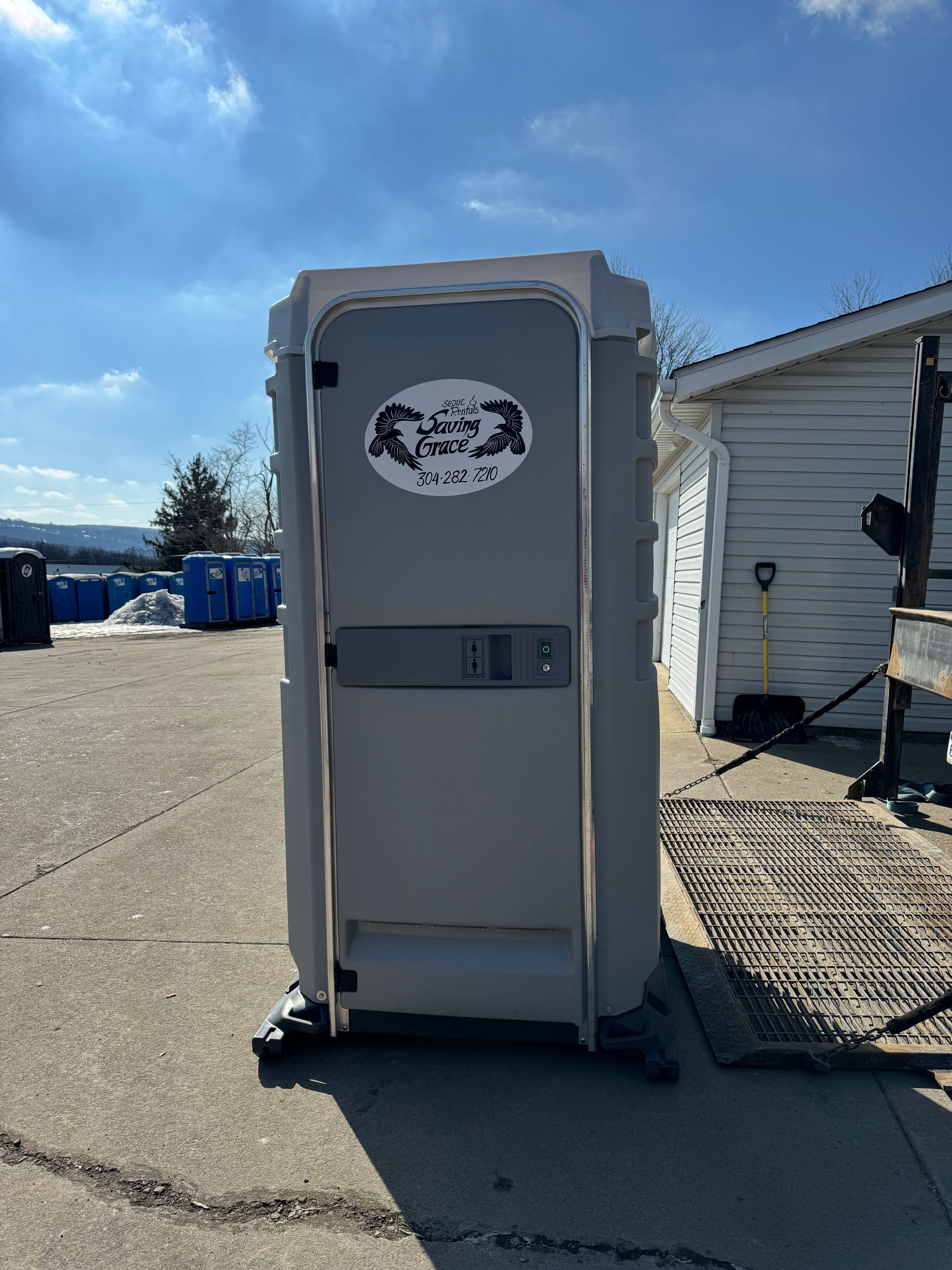 Gray portable toilet outside on a sunny paved lot beside a white building.