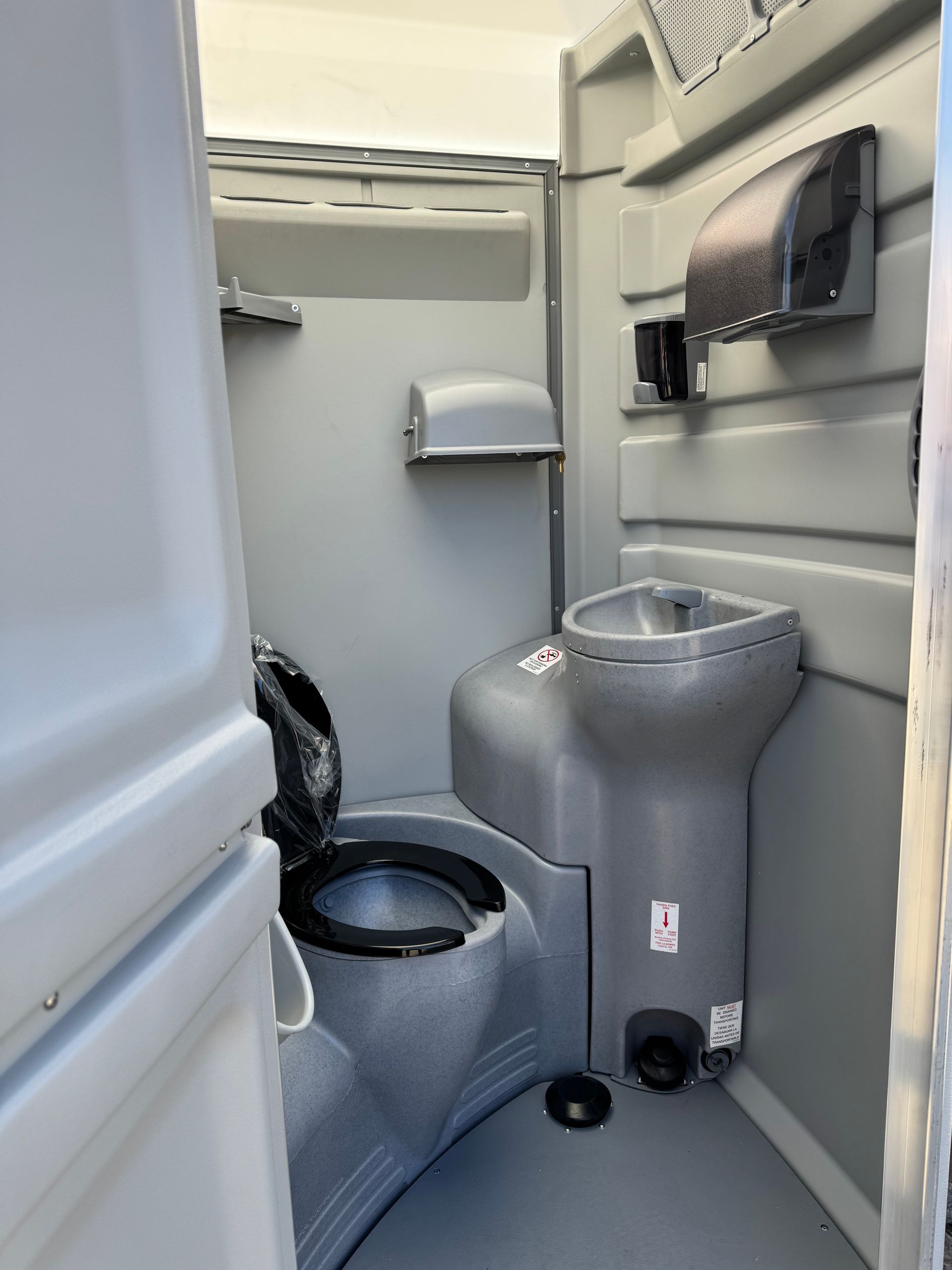 Portable toilet stall with gray toilet and sink, toilet paper dispenser, and wall-mounted hand sanitizer.