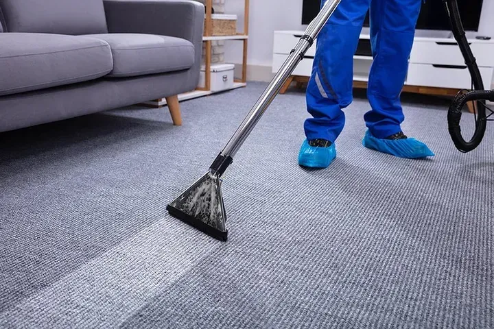 Person in blue uniform steam cleans gray carpet in a living room, leaving a clean path.