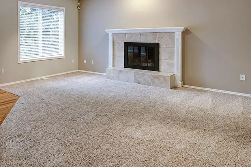 Empty living room with beige walls, carpet, fireplace, and window with shutters.