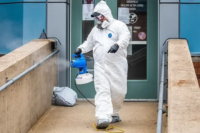 Person in hazmat suit disinfecting a building entrance with a sprayer.