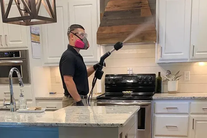 Person spraying kitchen range hood with a cleaning solution, wearing a mask and gloves.