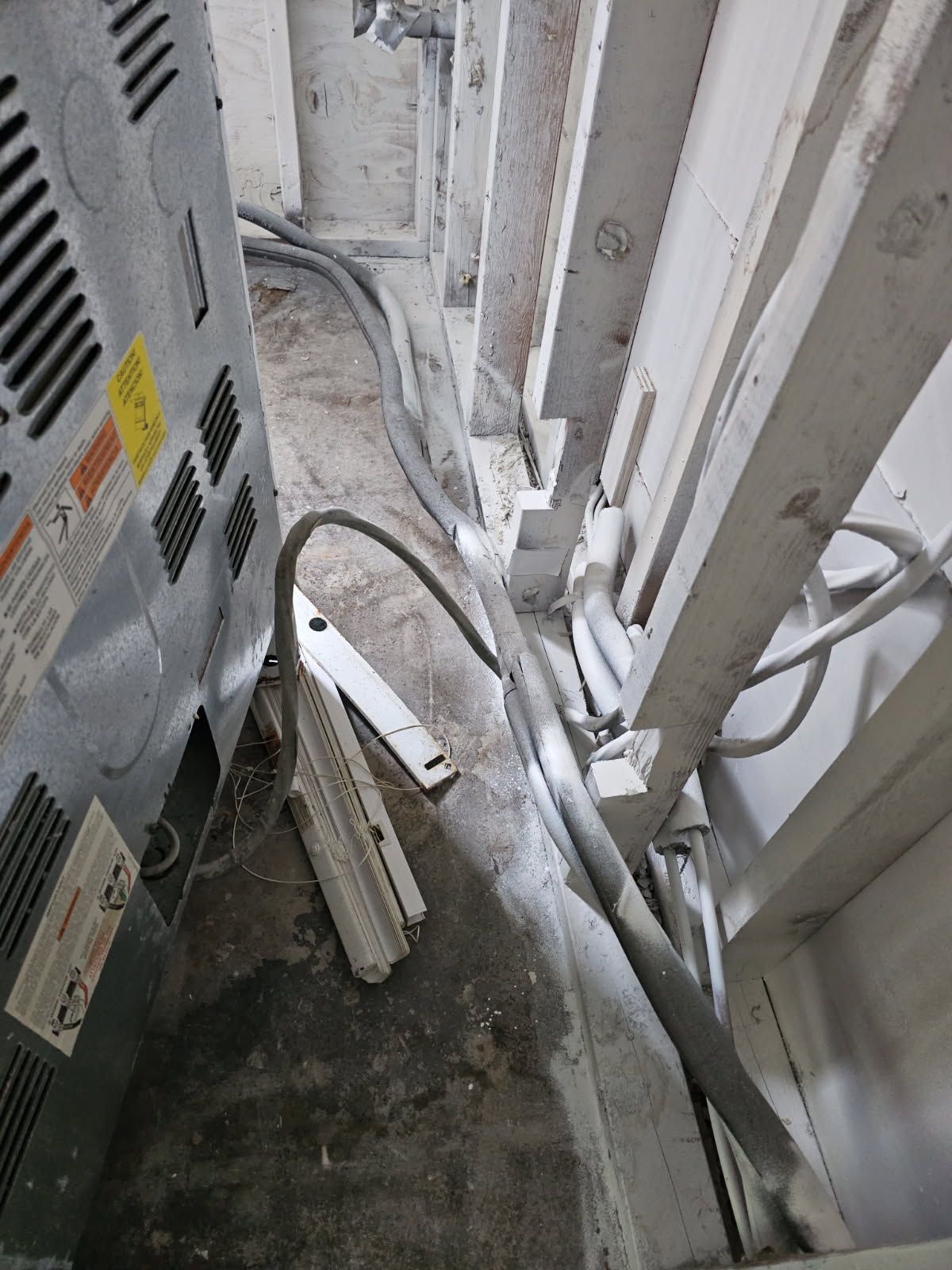 HVAC unit and electrical wiring in a narrow, unfinished space. Gray and white tones dominate, with debris on floor.