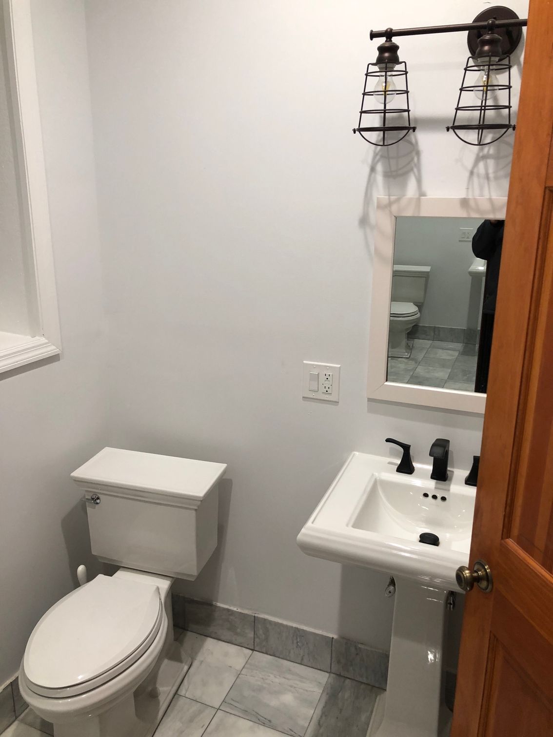 Bathroom with white toilet, pedestal sink, and caged light fixture. Gray tile floor.