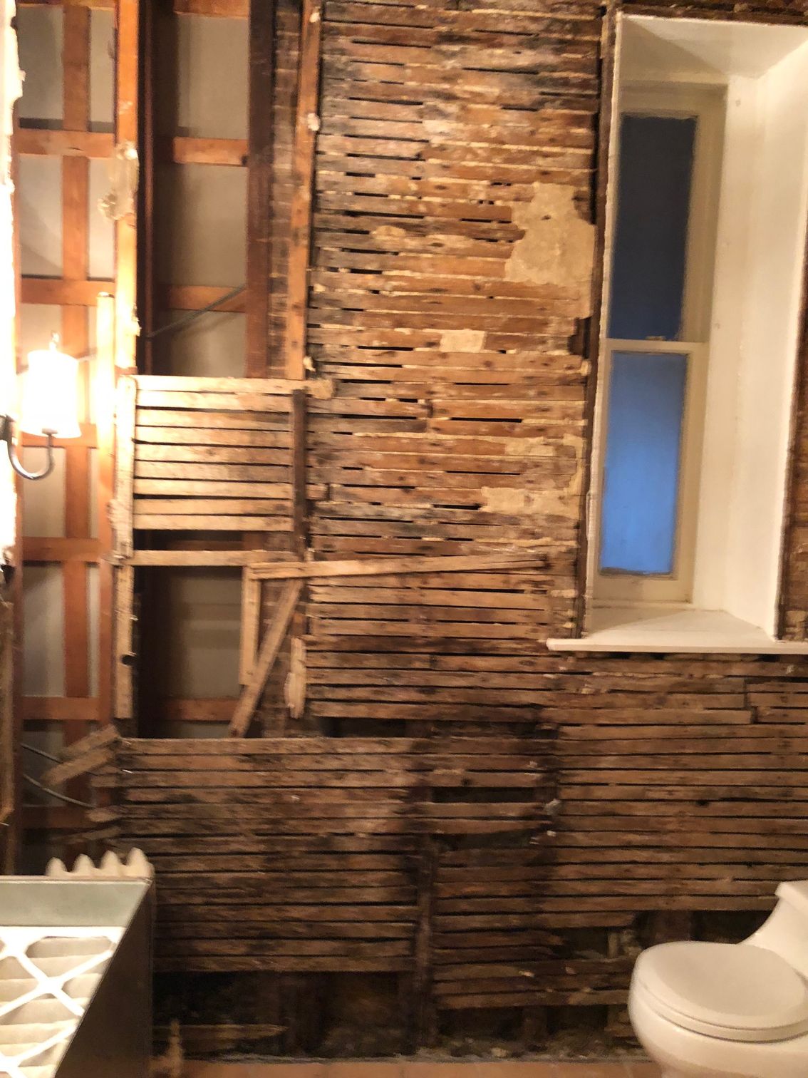 Bathroom wall with exposed wood lath, showing demolition during renovation. A window and toilet are visible.