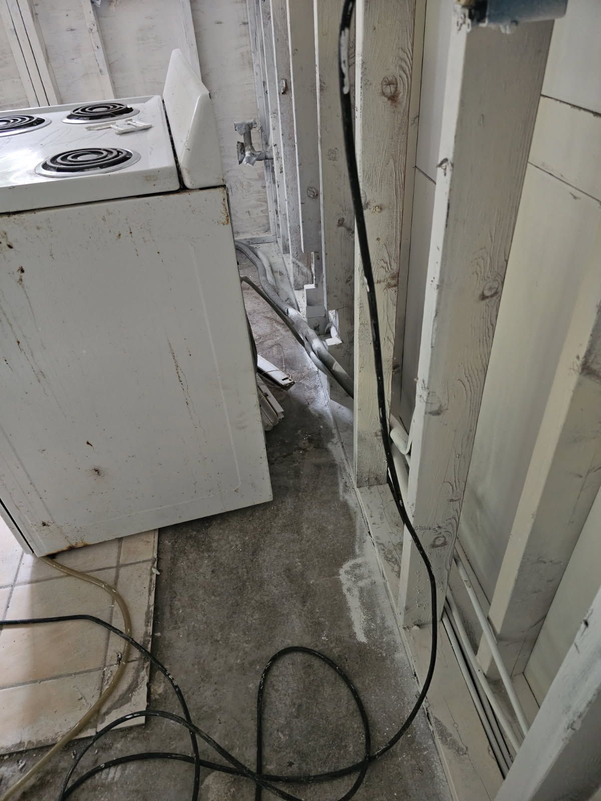 A messy kitchen with an old stove and wiring against a wall. Concrete floor, some water visible.