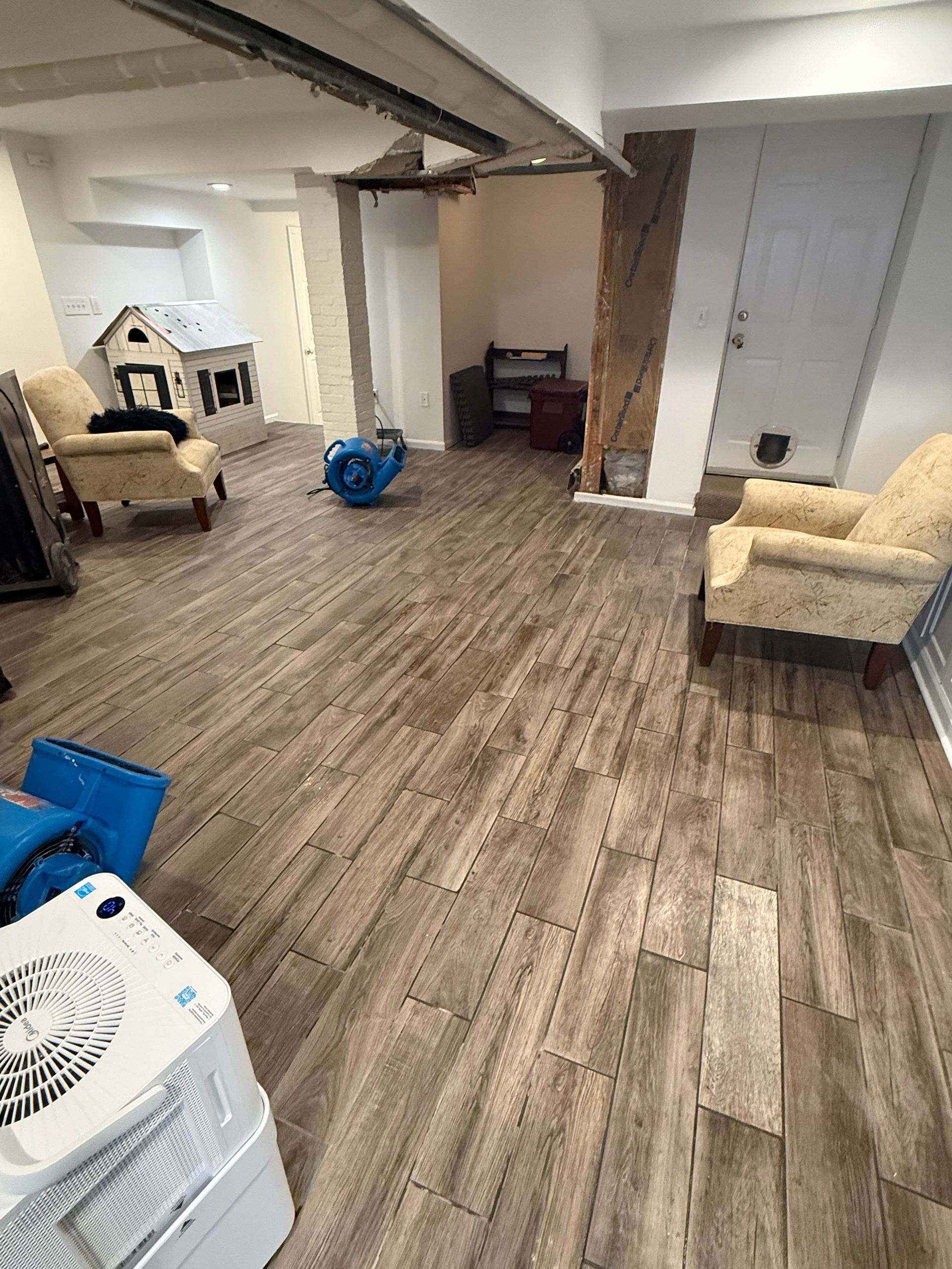 Basement with wood-look tile flooring, furniture, air movers, and a playhouse; neutral tones.