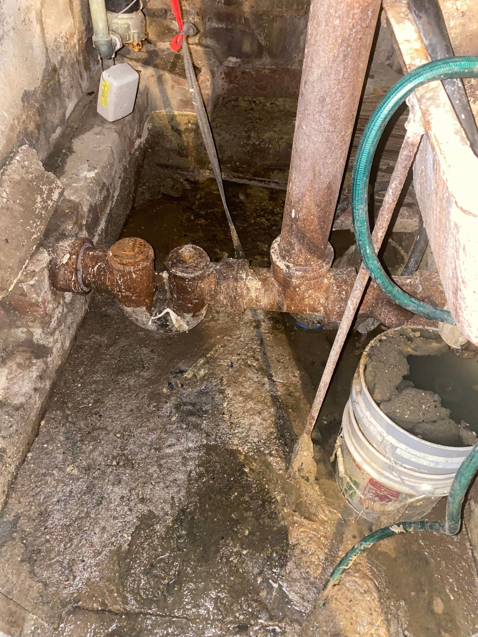 Flooded basement with rusty pipes, a bucket, and a hose; water covers the floor.