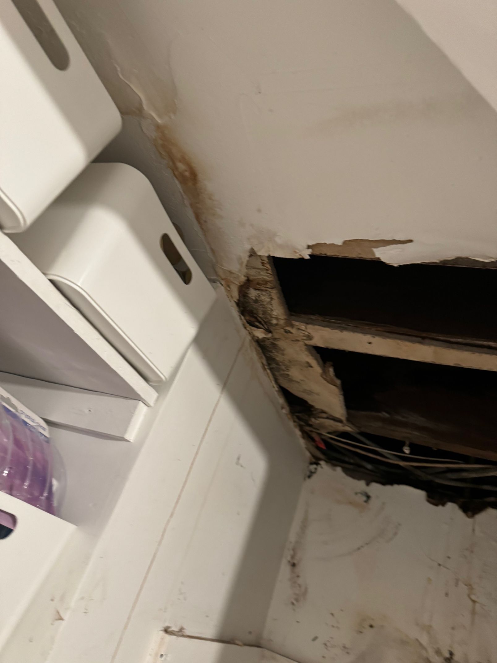 Damaged ceiling with a hole revealing wooden beams, likely due to water damage. White shelving unit on the left.