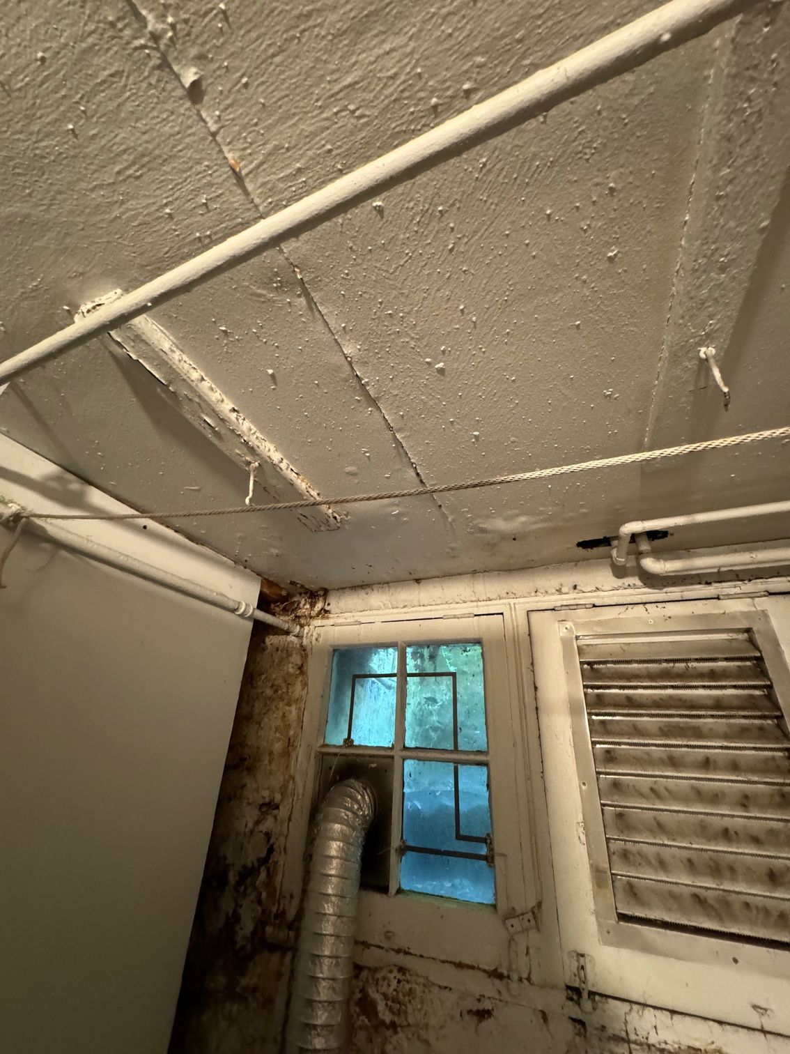 Basement ceiling with peeling paint, a small window, and exposed pipes.