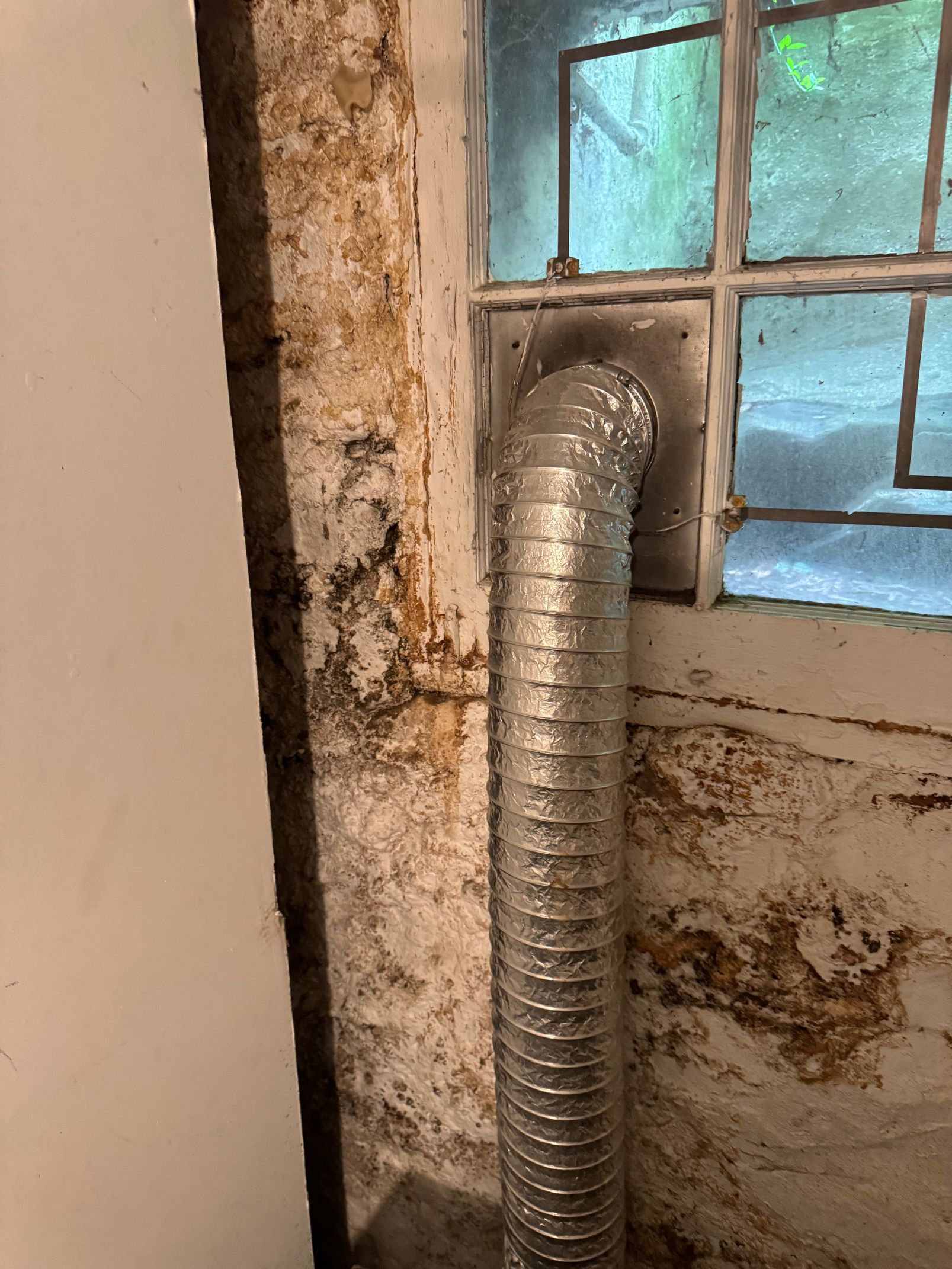 A flexible silver vent pipe connected to a wall, near a window with visible discoloration and possible mold.