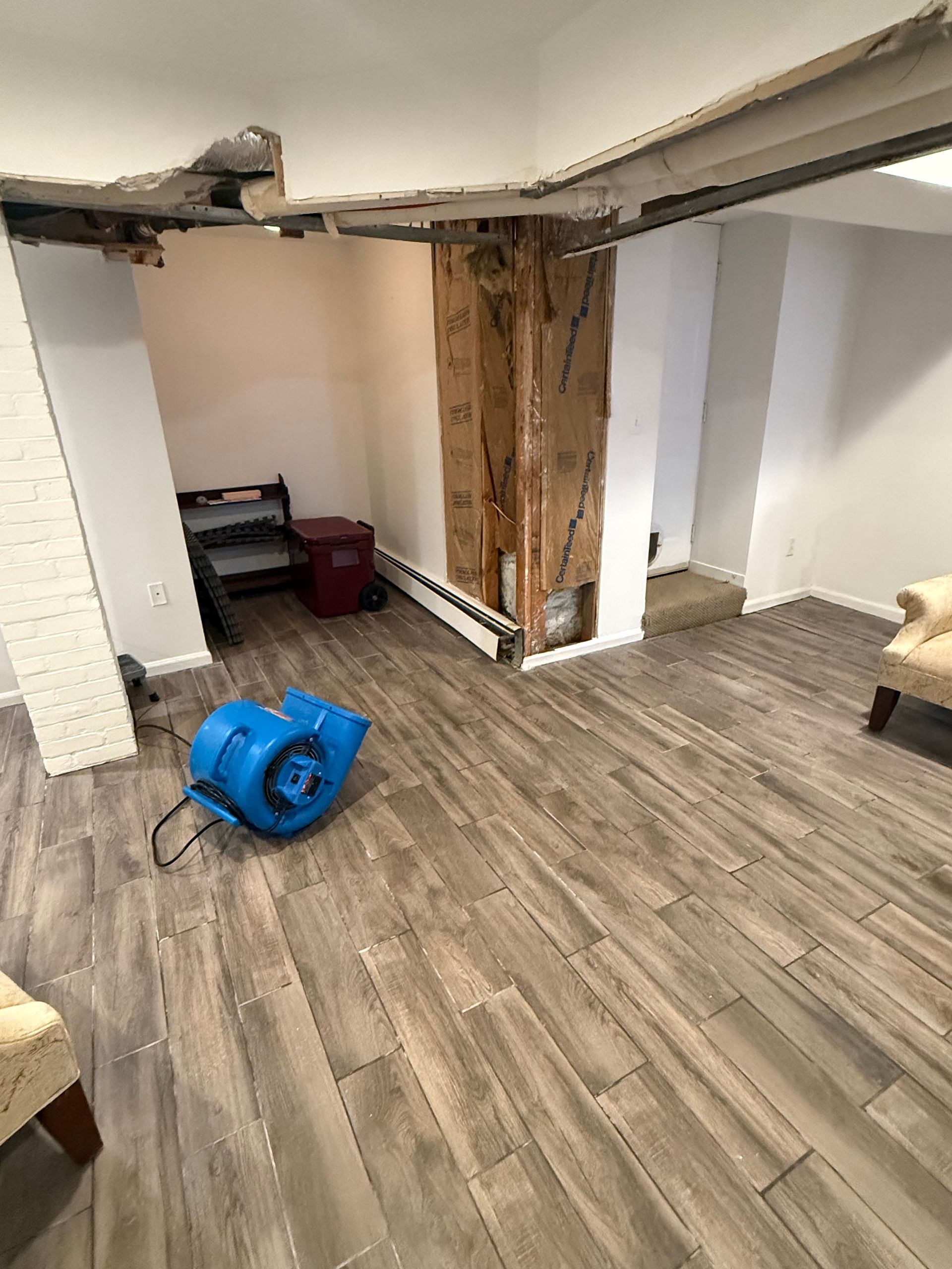 Room with damaged wall, drying fan, new wood-look flooring. Exposed ceiling beams and partially removed wall framing.