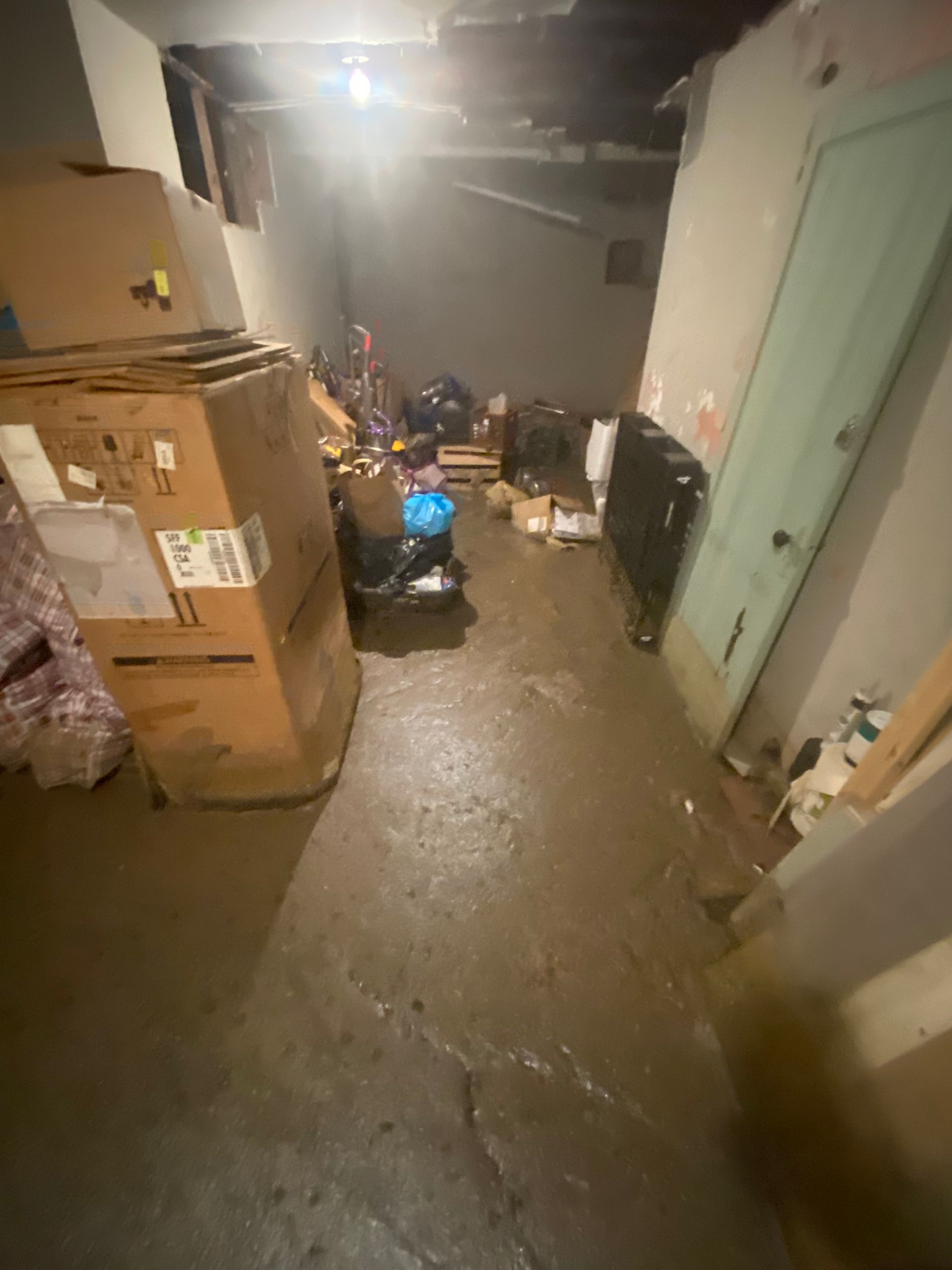 Cluttered basement with cardboard boxes, debris, and water on the floor; dim lighting.