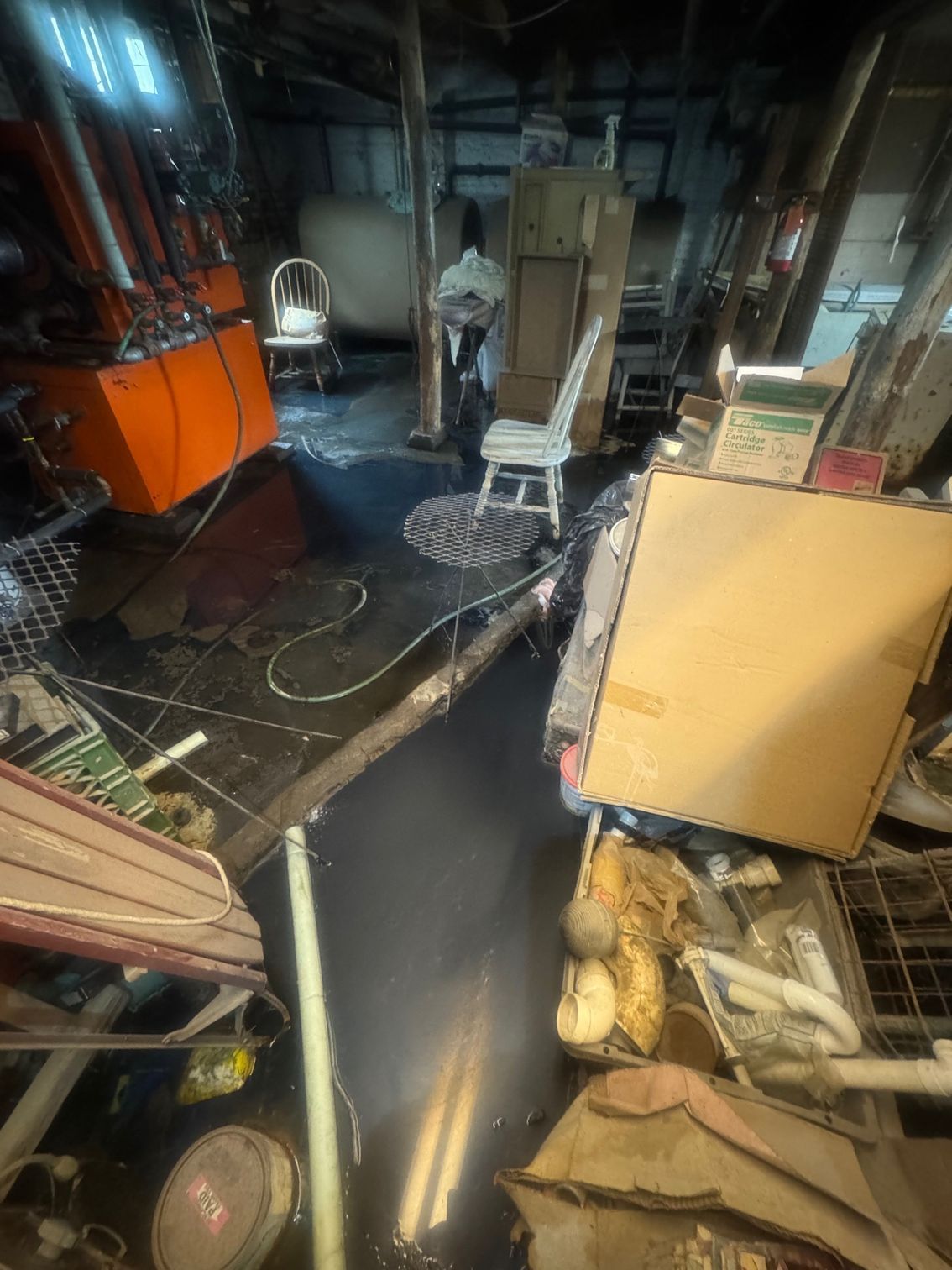 Messy basement with cluttered items, water on the floor, and white chairs.