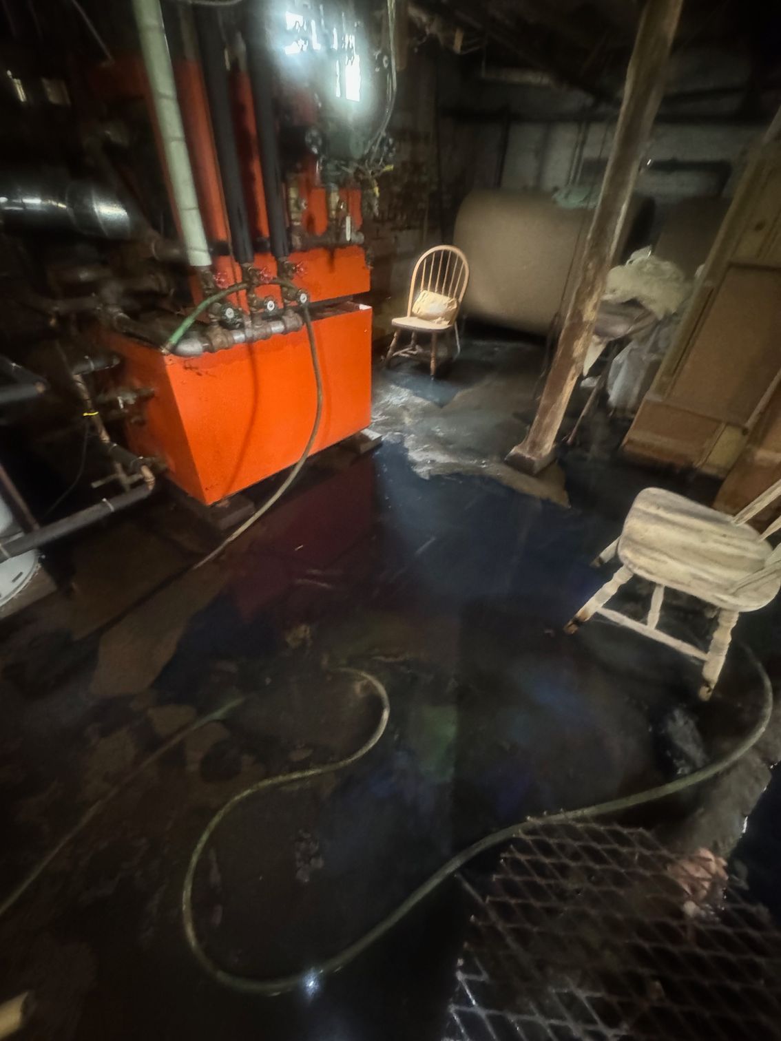 Orange industrial machine in a dark room with standing water on the floor, two chairs, and pipes.