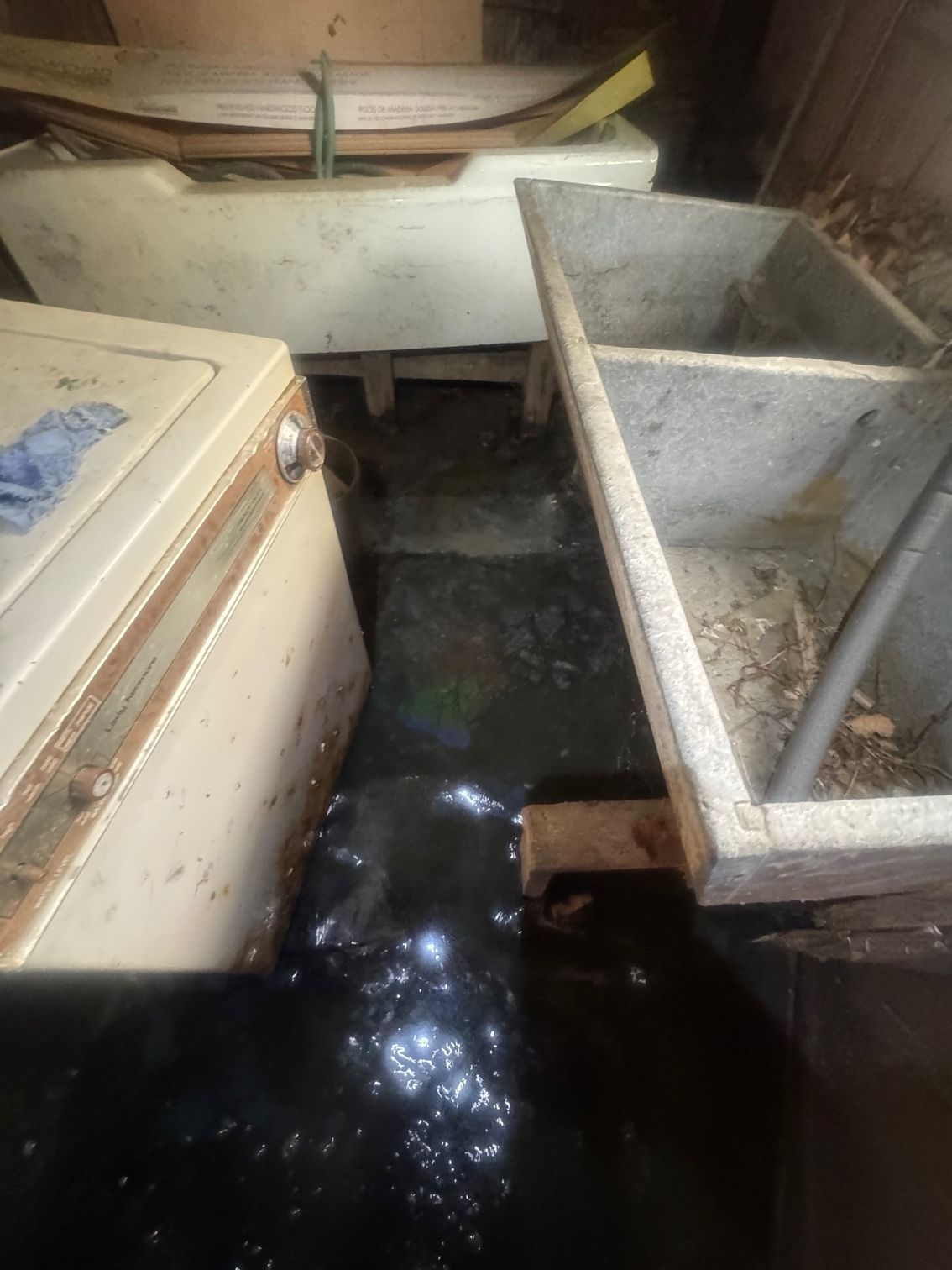 A flooded, dirty room with concrete washbasins and a chest-style appliance; water is dark and murky.