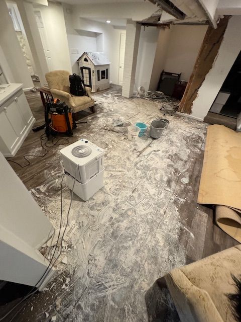 Basement with damaged flooring and construction debris; an air purifier and armchair are visible.