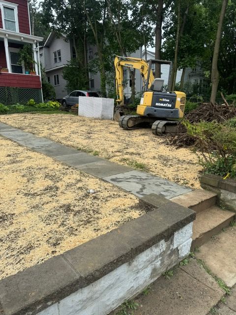 Excavator on a residential lot, with sidewalk and dirt. Houses and car in background.