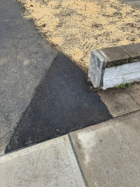 A triangular patch of fresh asphalt where sidewalk meets curb and curb meets gravel.