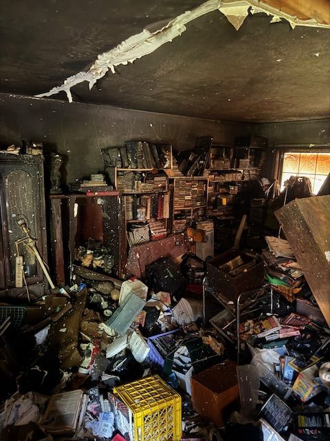 Messy room with smoke damage, piled with debris, shelves, and cluttered furniture.