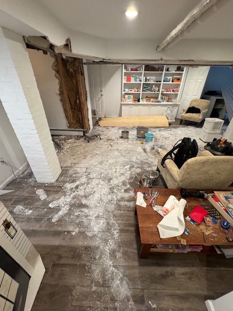 Room with debris on the floor, damaged wall, and exposed wooden beam.  Bookshelves and furniture present.