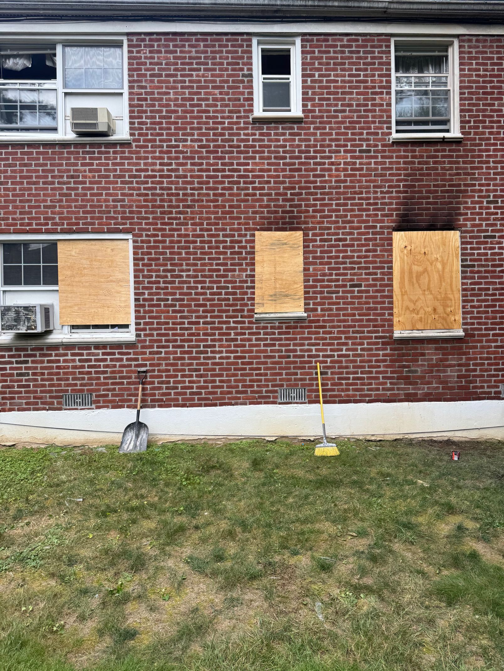 Brick building with boarded-up windows and tools on the ground; grass in foreground.