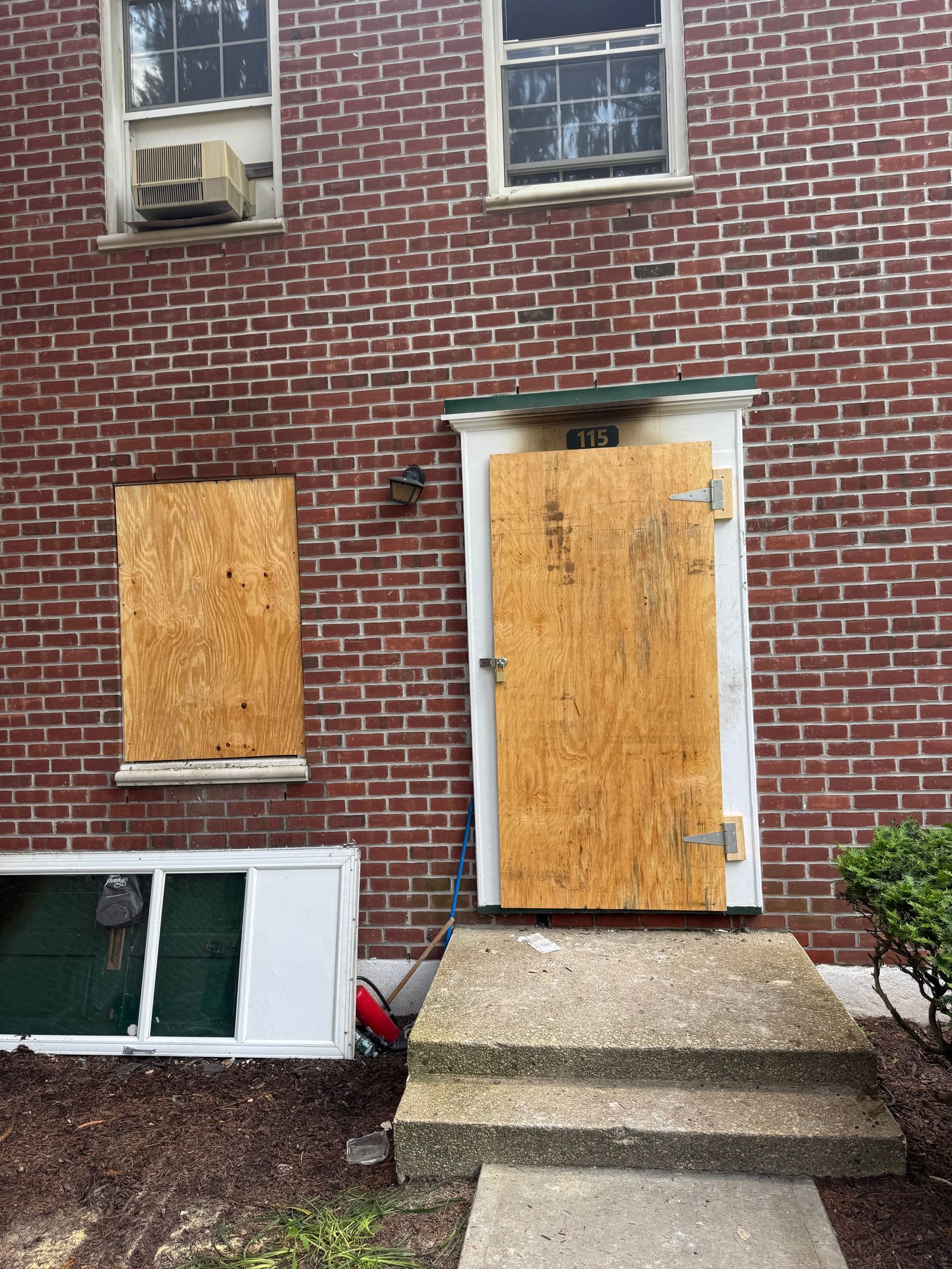 Exterior of brick building with boarded-up door and window; concrete steps, overgrown bush.