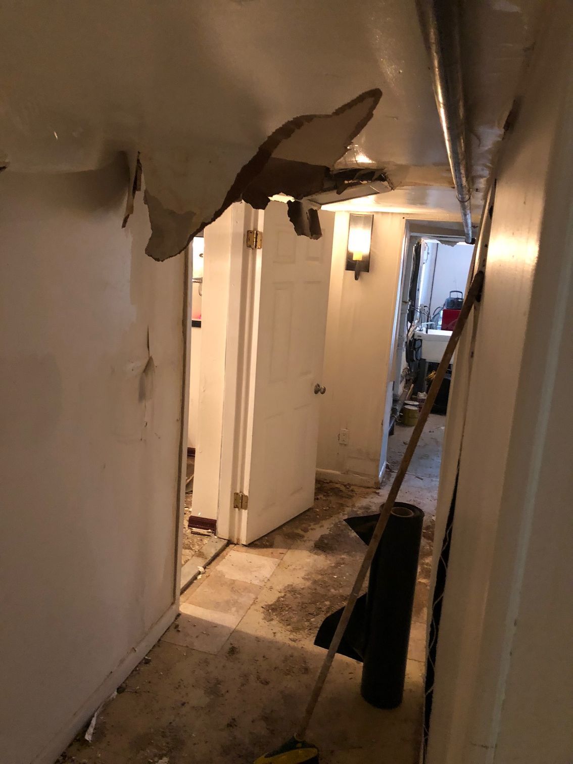 Damaged ceiling in a narrow hallway with debris on the floor and a broom propped against the wall.