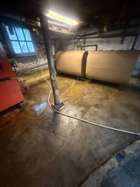 Basement with a concrete floor, oil tank, support beam, and water hose. Low lighting.