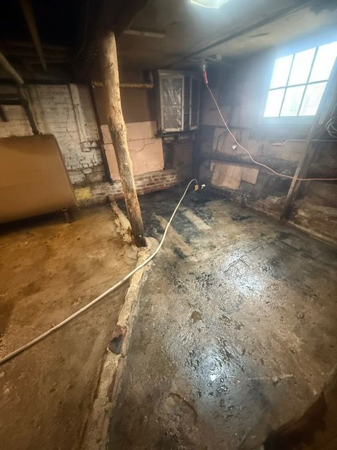 A dark, damp basement with concrete floor, wooden beams, and a small window.