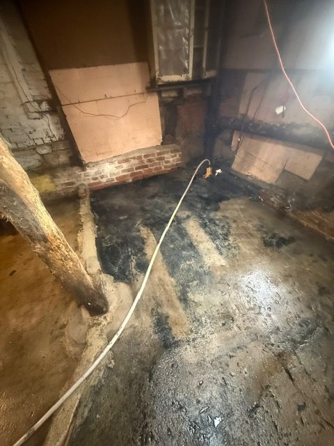 Dark, damp basement corner with exposed beams, brick, and pooling water. Electrical cord lies across the floor.