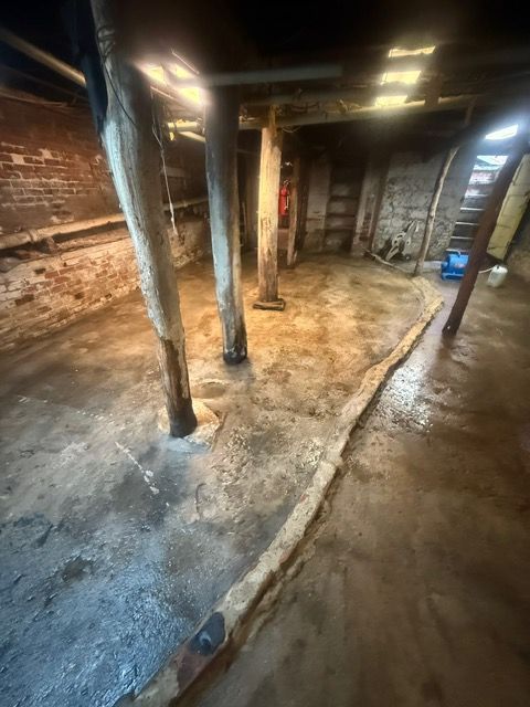 Old basement with concrete floor, wooden support beams, brick walls, and stairs.