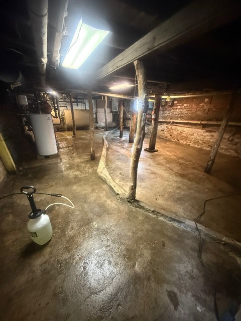 Wet basement with exposed beams and support posts, lit by a fluorescent light. A sprayer is on the floor.