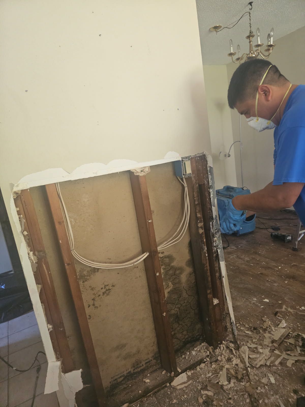 Man in mask working on damaged wall, visible wiring and mold, debris on floor.