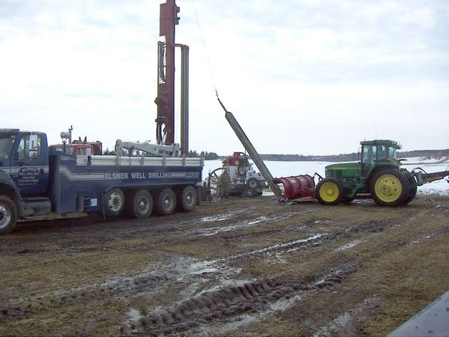 A blue utility truck with a drilling mast sits on a muddy field next to a green tractor pulling a red agricultural tool.