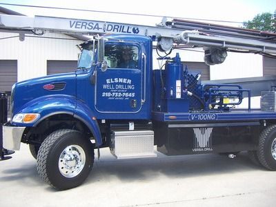 A blue Versa-Drill well drilling truck parked in front of a white building.