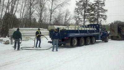 Three people in winter clothing working with hoses and a large dark blue truck on a snow-covered field.