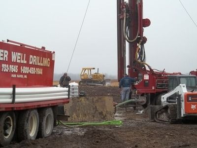 A red well drilling truck and a drilling rig operating on a muddy construction site, with a yellow tractor in the distance.