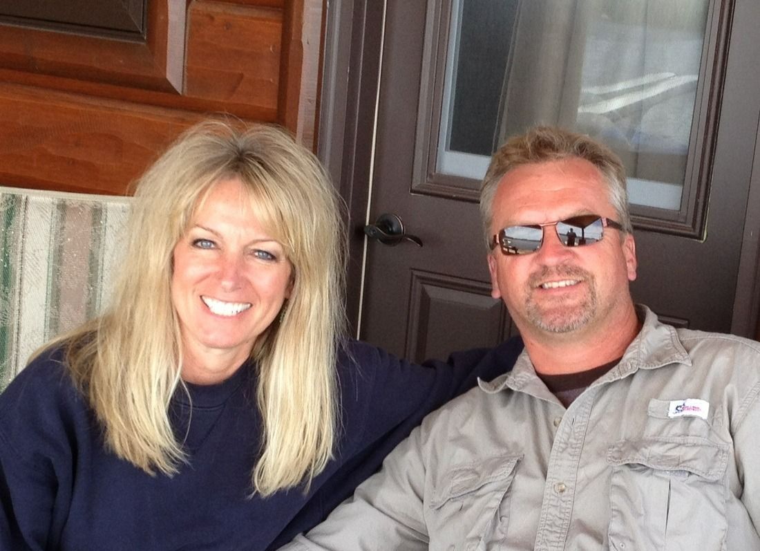 A smiling woman and a man in sunglasses sitting together outdoors in front of a wood-paneled wall and a door.