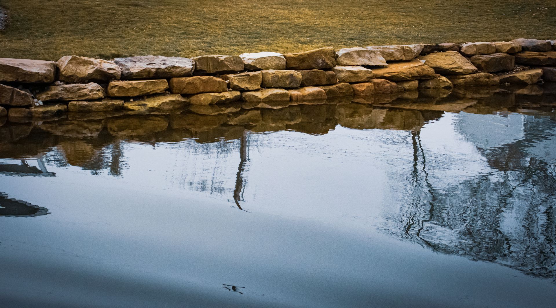 A stone wall is reflected in the water of a pond.