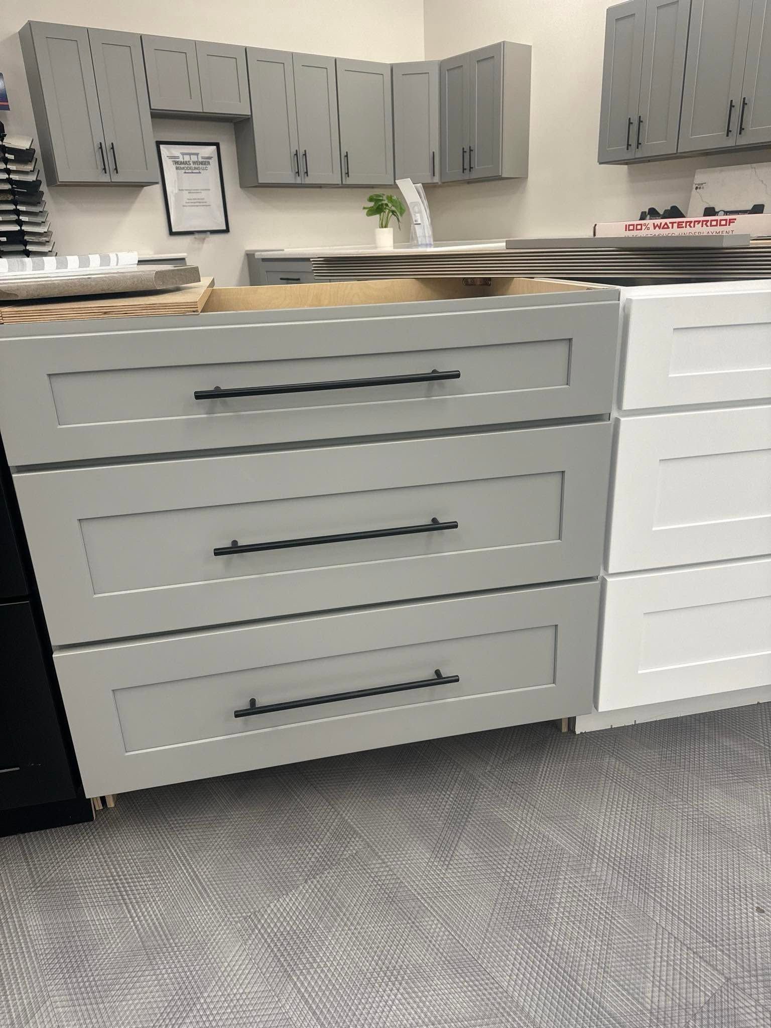 Kitchen showroom with gray cabinets, white drawers, and a speckled countertop on a gray carpet.